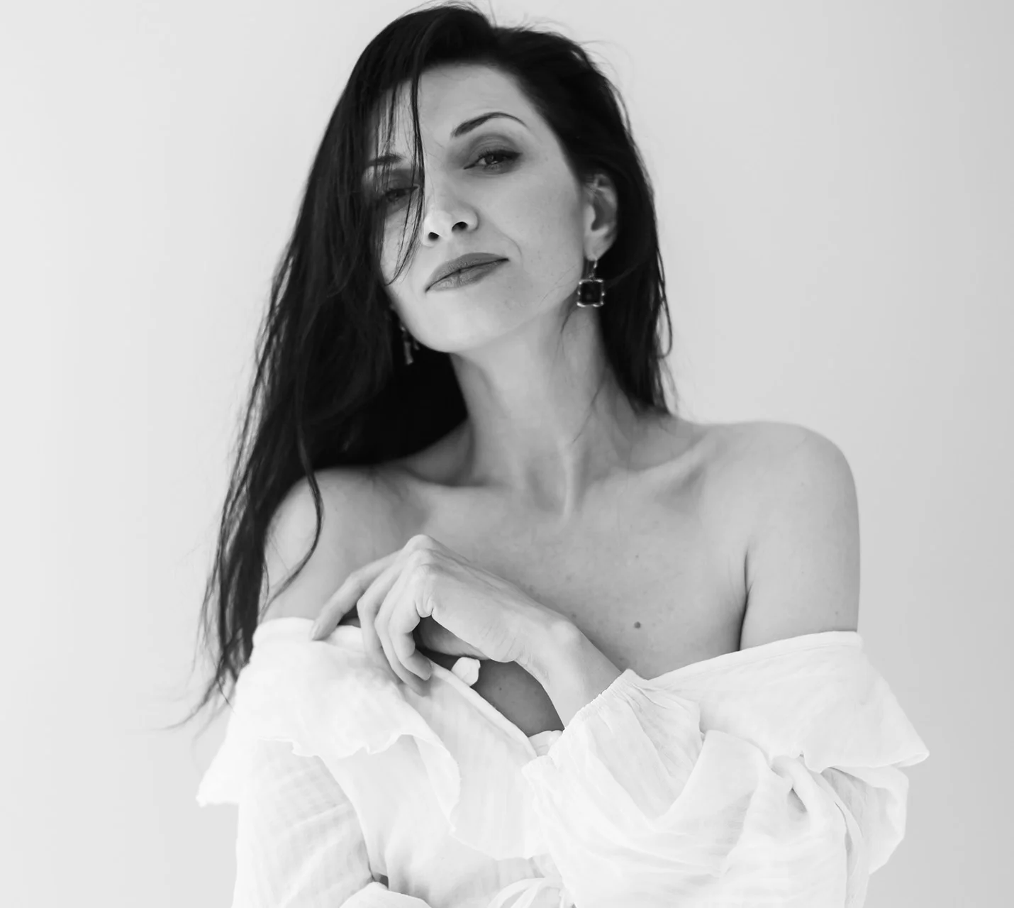 A woman with long dark hair poses confidently, wearing an off-the-shoulder white blouse, against a light background. - Breast Cancer Reconstruction in West Village, New York, NY