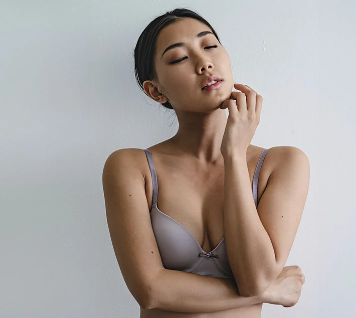 A woman in a light purple bra stands against a plain wall, with her eyes closed and a serene expression, hand on her chin. - Breast Implant Capsular Contracture in West Village, New York, NY