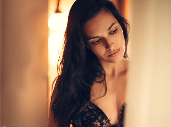A woman with long dark hair gazes down thoughtfully, wearing a delicate black lace garment against a softly lit background. - Breast Reduction PP in West Village, New York, NY