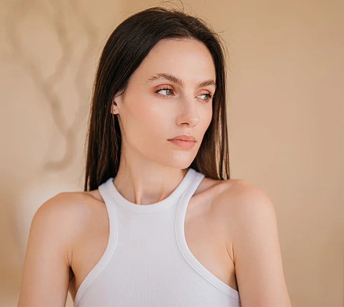 A woman with long dark hair gazes thoughtfully off-camera, wearing a sleeveless white top against a soft background. - Brow Lift in West Village, New York, NY