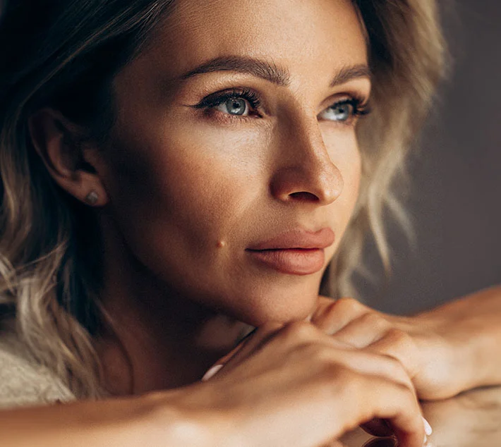 A thoughtful woman gazes to the side, resting her chin on her hands, with soft lighting highlighting her features. - Brow Lift in West Village, New York, NY