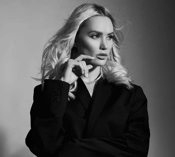 A striking black-and-white portrait of a woman with long, wavy blonde hair, wearing a stylish black blazer and posing thoughtfully. - Buccal Fat Removal in West Village, New York, NY