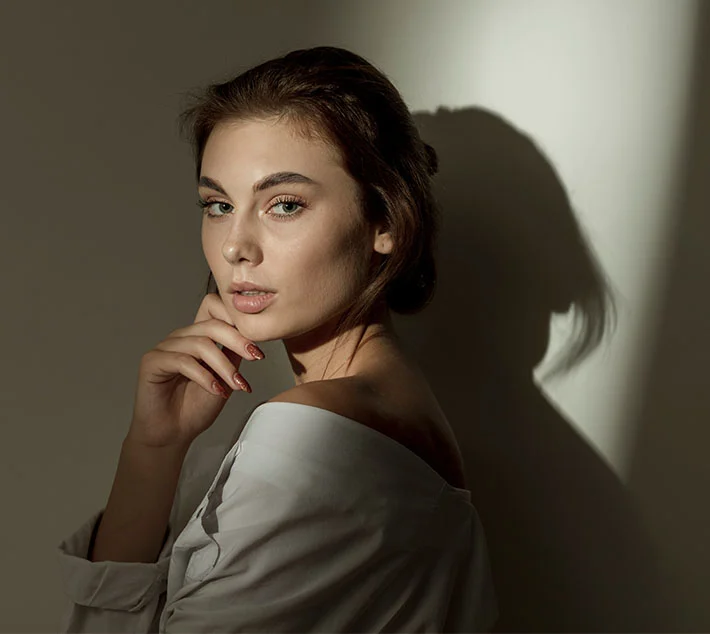 A woman with long brown hair gazes at the camera, gently touching her face, with soft light casting a shadow behind her. - Buccal Fat Removal in West Village, New York, NY