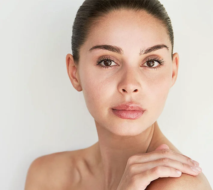 A close-up of a woman with smooth skin, dark hair pulled back, and a natural expression, softly touching her shoulder. - Chin Augmentation in West Village, New York, NY