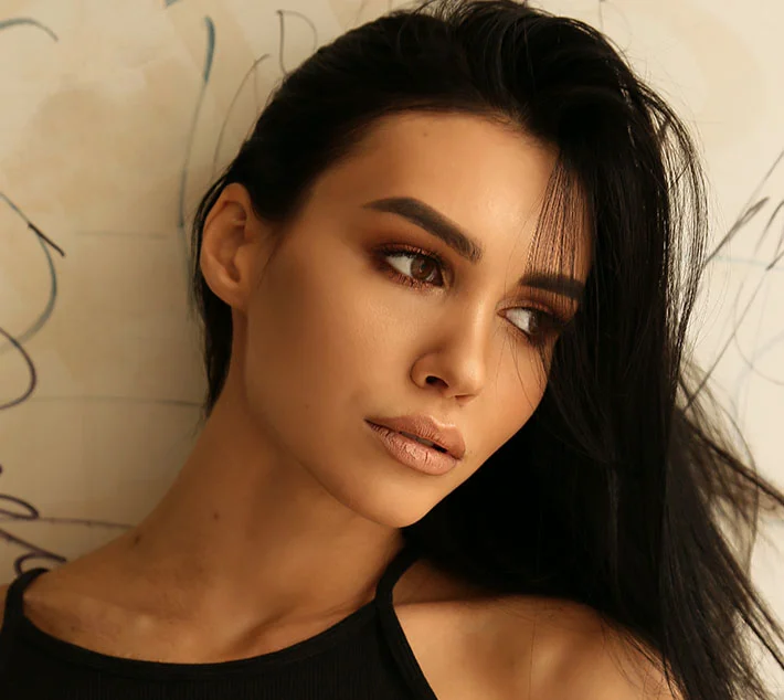 A woman with long black hair poses against a lightly textured wall, showcasing a subtle makeup look and a pensive expression. - Chin Augmentation in West Village, New York, NY