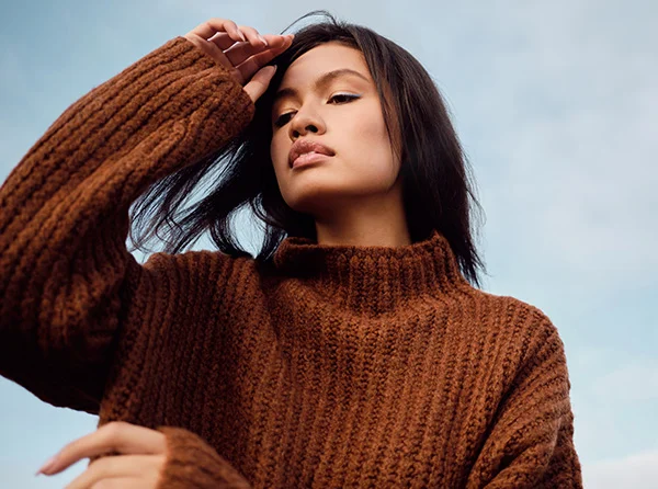 A young woman with shoulder-length dark hair, wearing a brown chunky sweater, gazes thoughtfully against a blue sky backdrop. - Chin Augmentation in West Village, New York, NY
