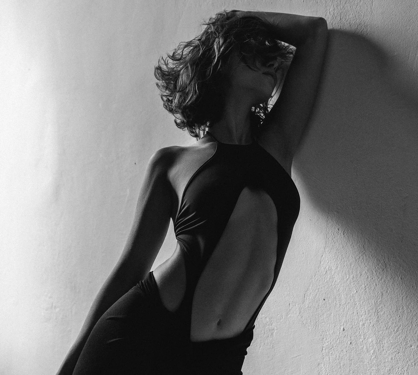 A silhouette of a woman in a black cut-out dress poses against a textured wall, her hair tousled with a dramatic flair. - Drainless Tummy Tuck in West Village, New York, NY