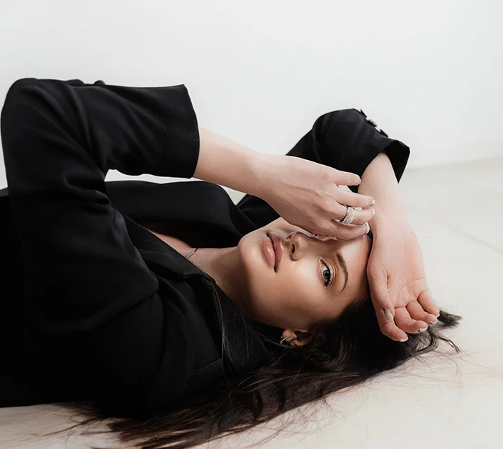 A woman with long hair lies on a floor, wearing a black blazer, resting her chin on her hand while gazing at the camera. - Eyelid Surgery in West Village, New York, NY
