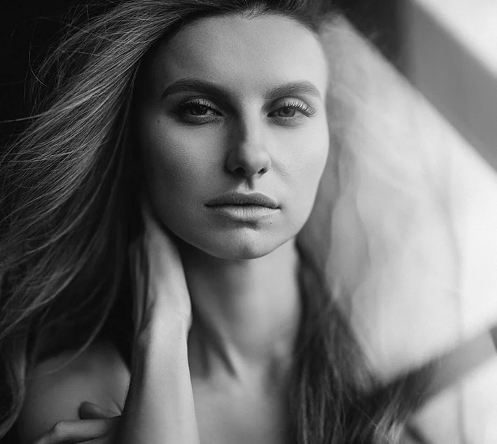 A close-up black and white portrait of a woman with long hair, gazing intensely at the camera, with soft lighting. - Eyelid Surgery in West Village, New York, NY