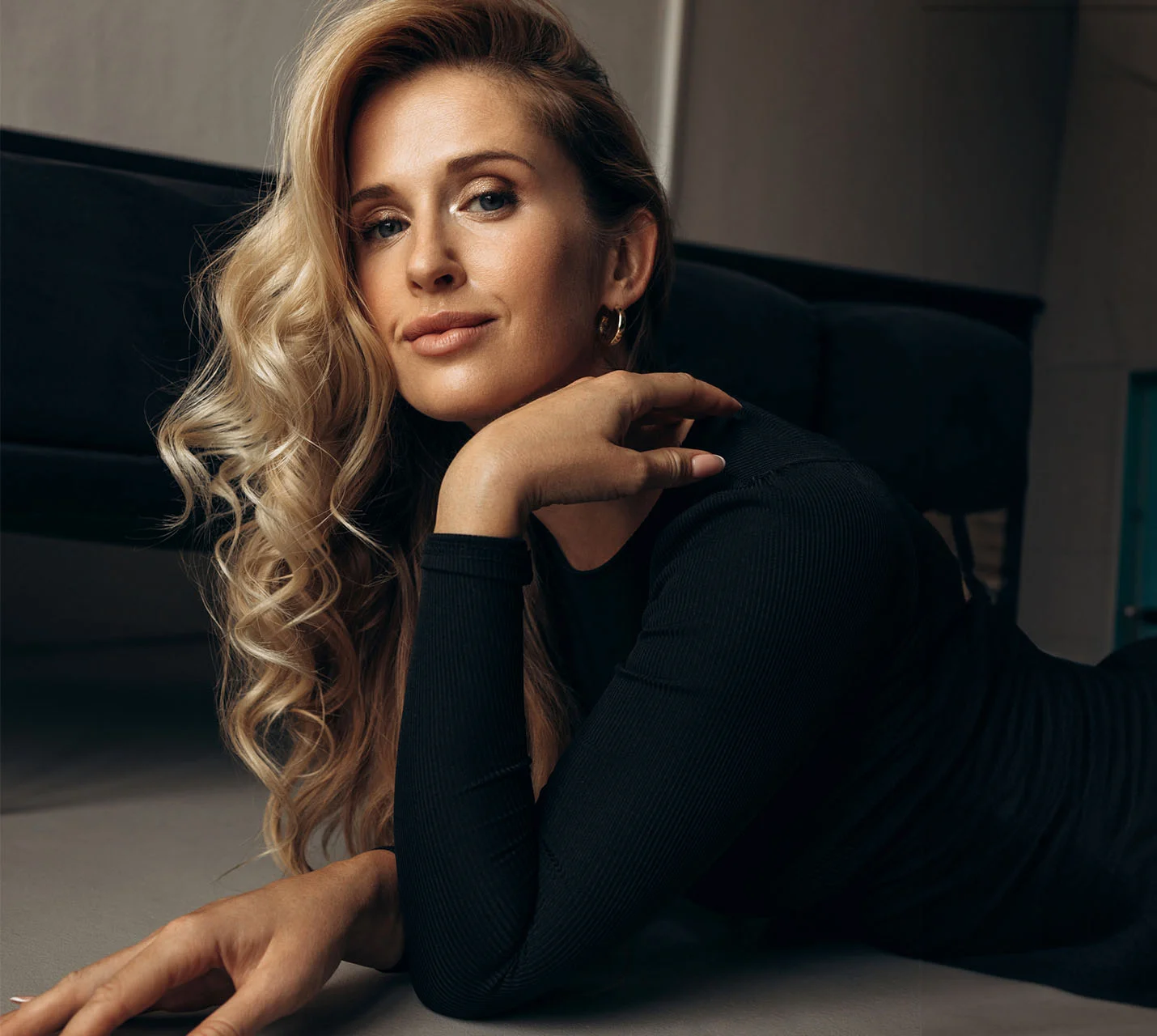 A woman with long, wavy blonde hair lies on the floor, resting her chin on her hand, smiling softly at the camera. - Eyelid Surgery in West Village, New York, NY