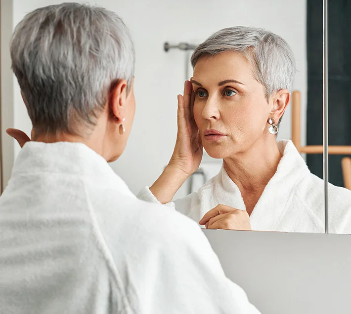 A woman with short gray hair in a white robe touches her face while looking into a bathroom mirror, contemplating her reflection. - Face Lift in West Village, New York, NY