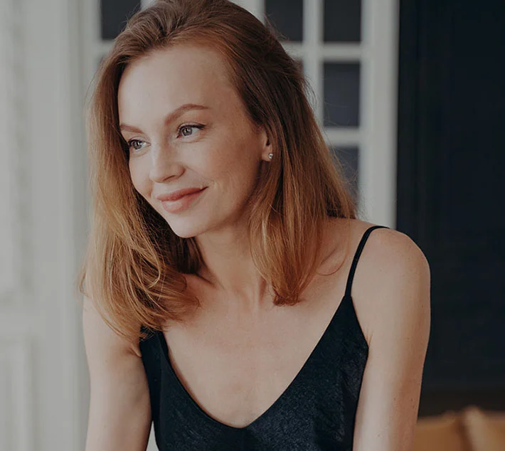 A woman with long, light brown hair smiles softly, wearing a black tank top, against a softly lit background. - Face Lift in West Village, New York, NY