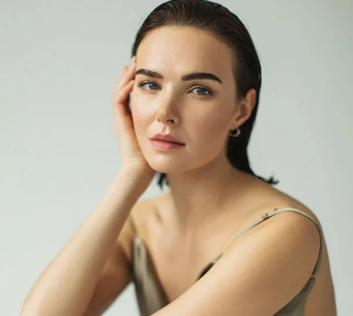 A young woman gazes softly at the camera, with her hand resting against her cheek, wearing a simple, elegant outfit. - Facial Grafting Breast Augmentation in West Village, New York, NY