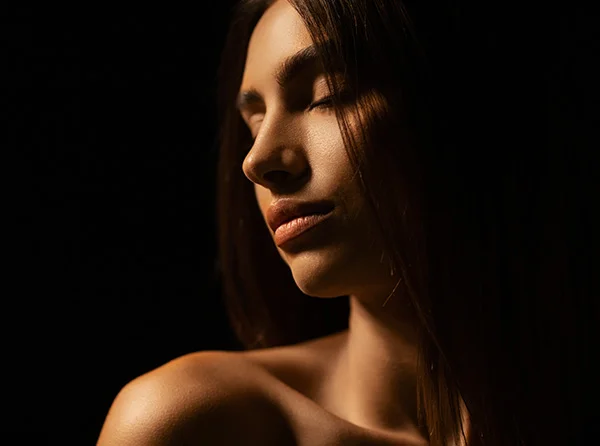 A close-up of a woman with closed eyes, softly illuminated by warm light against a dark background, showcasing serene beauty. - Facial Fat Grafting in West Village, New York, NY
