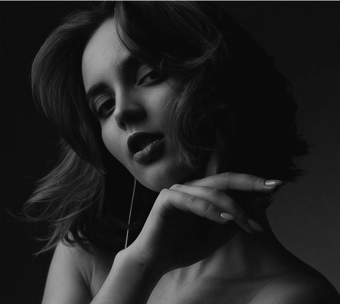 A dramatic black and white portrait of a woman with wavy hair, softly resting her chin on her hand, showcasing bold lips. - Facial Grafting Breast Augmentation in West Village, New York, NY