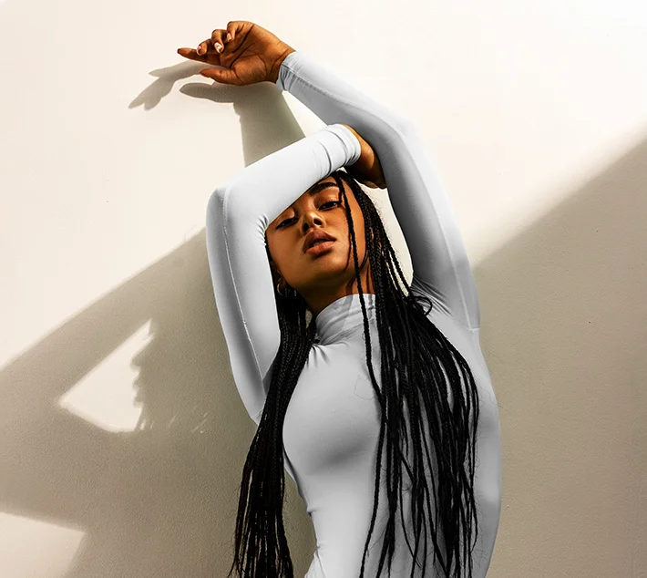 A person in a fitted white outfit poses against a light wall, with long braided hair cascading down, creating dramatic shadows. - Fat Grafting Breast Augmentation in West Village, New York, NY