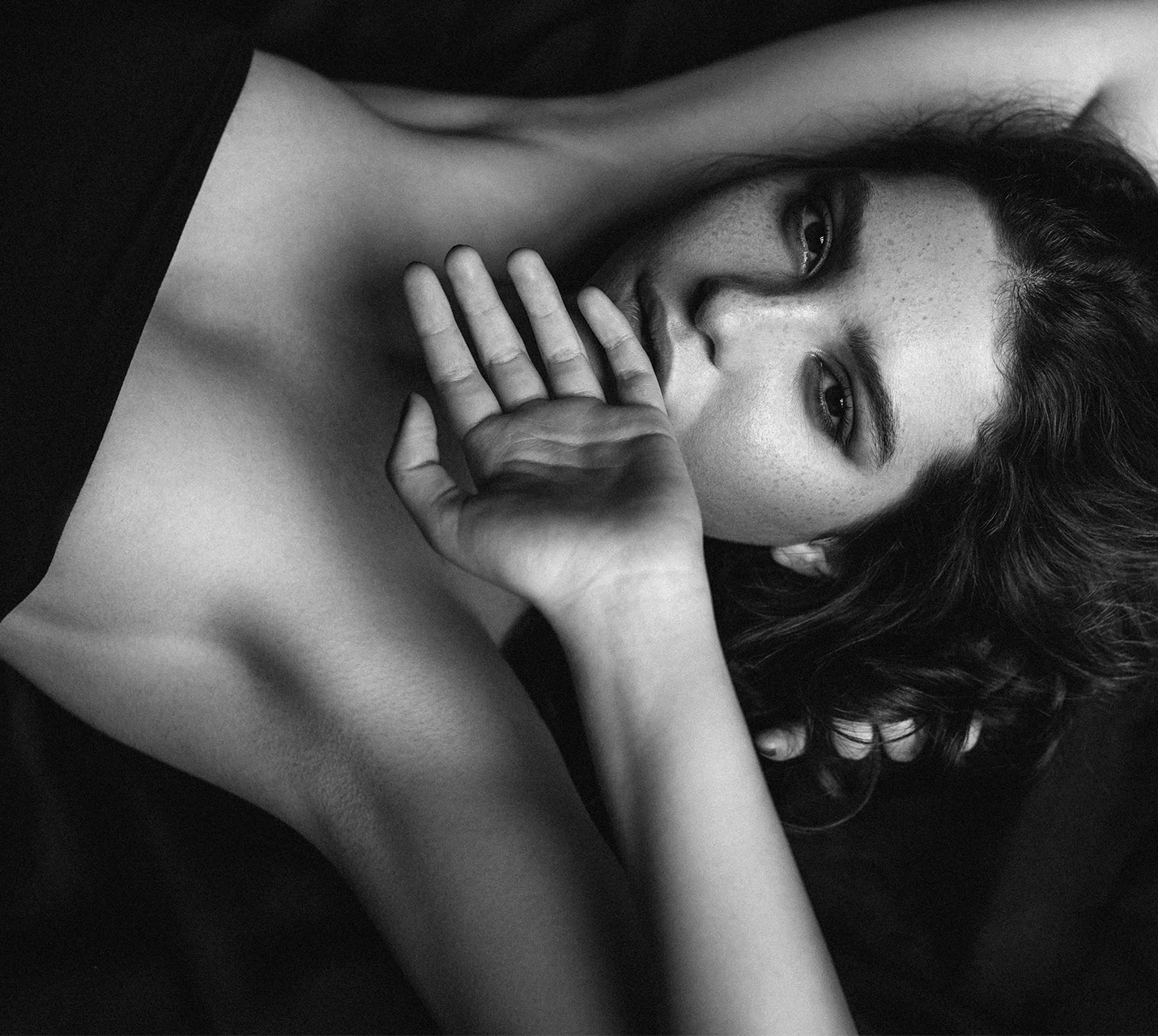 A close-up of a person with curly hair, resting on a dark surface, gazing intently while covering part of their face with a hand. - Fat Grafting Breast Augmentation in West Village, New York, NY