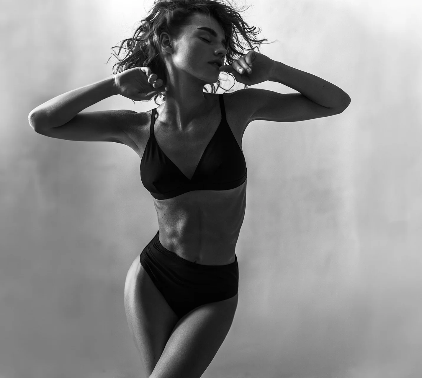 A woman poses elegantly in black swimwear, her arms raised, showcasing a confident and graceful silhouette against a soft backdrop. - High Definition Liposuction in West Village, New York, NY