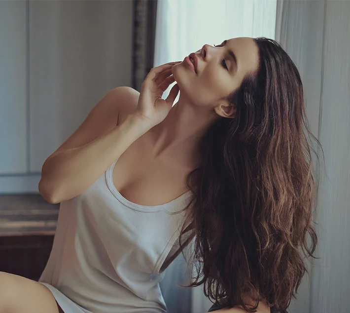 A woman with long hair poses gracefully, touching her neck with one hand, looking thoughtfully towards the window. - Internal Bra in West Village, New York, NY