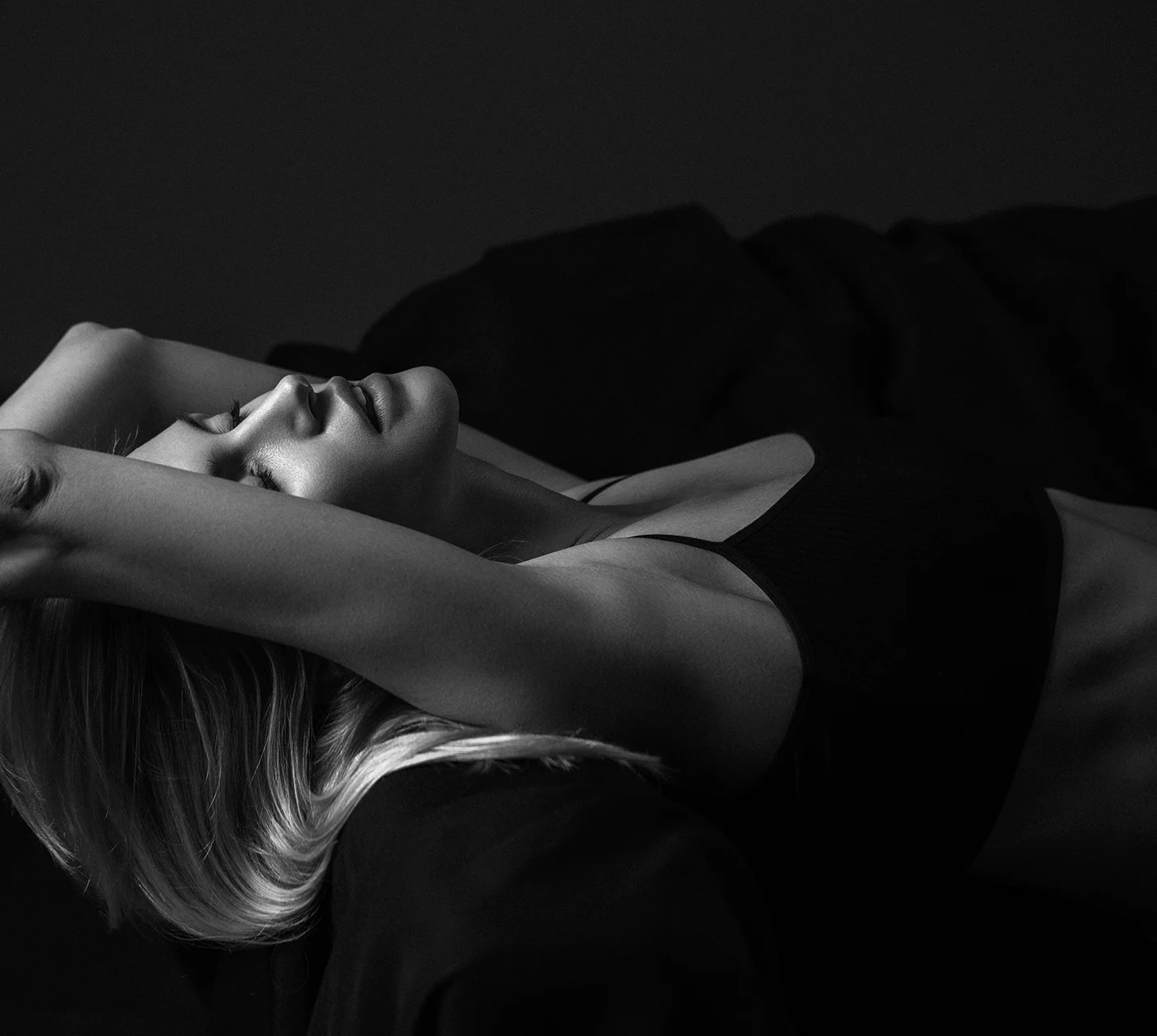 A woman with blonde hair lies back on a dark surface, arms lifted overhead, in a black and white artistic photograph. - Internal Bra in West Village, New York, NY
