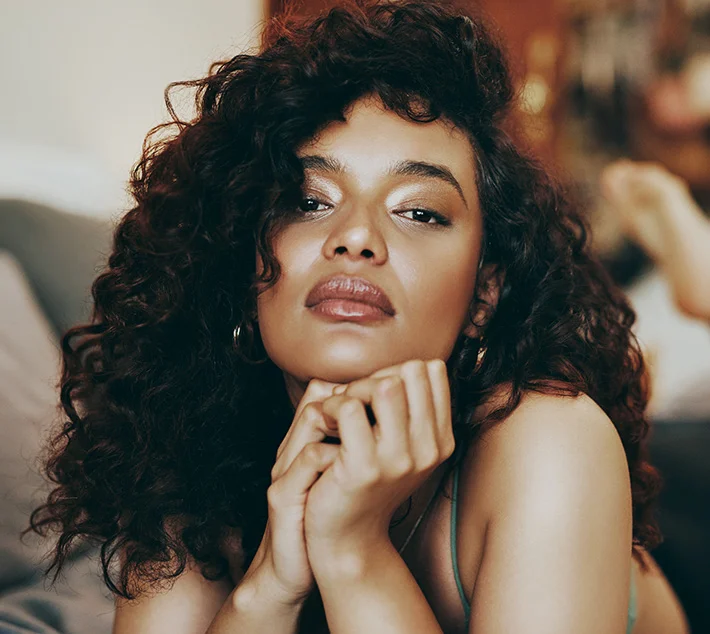 A woman with curly hair gazes thoughtfully at the camera, resting her chin on her hands in a cozy indoor setting. - Lip Lift in West Village, New York, NY