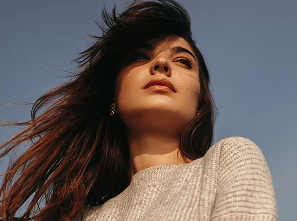 A young woman with flowy hair gazes upward against a clear sky, wearing a gray sweater and a serene expression. - Neck and Chin Liposuction in West Village, New York, NY