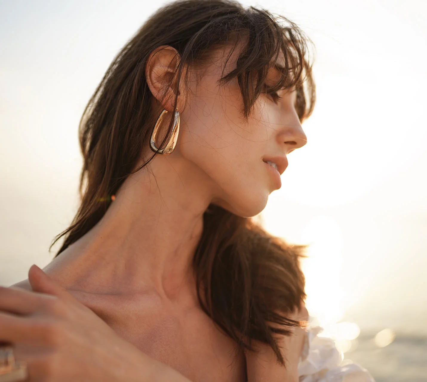 A profile of a woman with long, wavy hair wearing large gold earrings, softly illuminated by a sunset at the beach. - Neck Liposuction in West Village, New York, NY