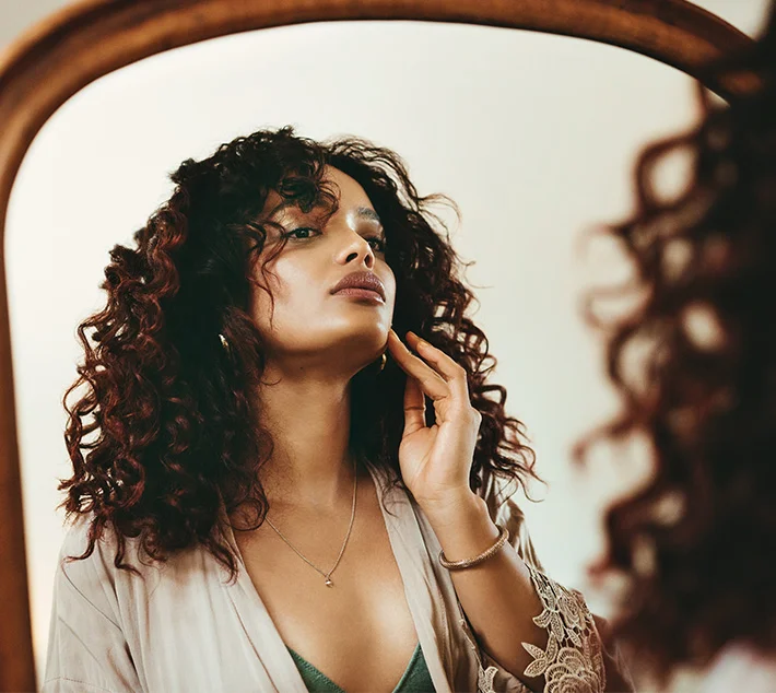A woman with curly hair softly examines her reflection in a vintage mirror, dressed in a light robe and necklace. - Neck Lift in West Village, New York, NY