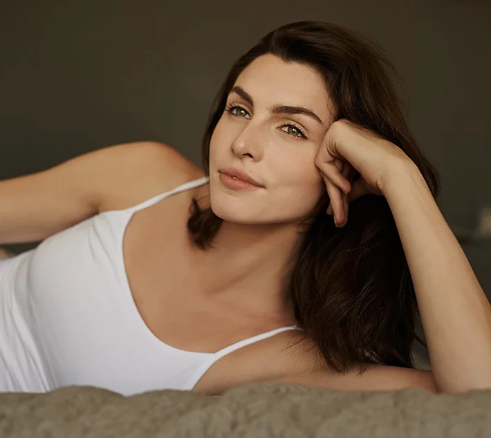 A woman with long brown hair rests on a bed, smiling softly while propping her head on her hand against a dim backdrop. - Rhinoplasty in West Village, New York, NY