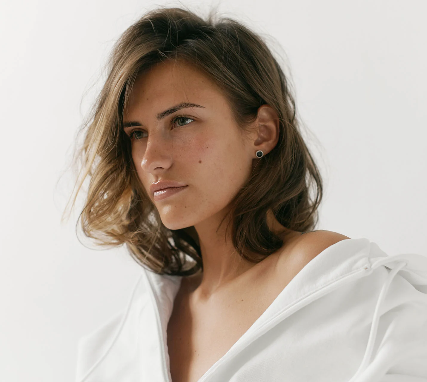 A close-up portrait of a woman with tousled hair, wearing a white oversized hoodie, gazing thoughtfully into the distance. - Rhinoplasty in West Village, New York, NY