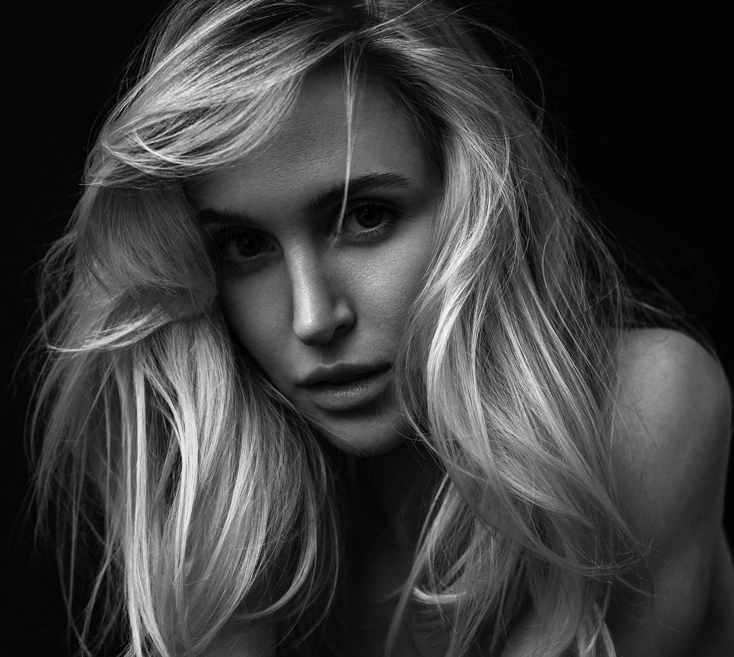 A close-up black-and-white portrait of a woman with long, wavy blonde hair and a serious expression against a dark background. - Rhinoplasty in West Village, New York, NY
