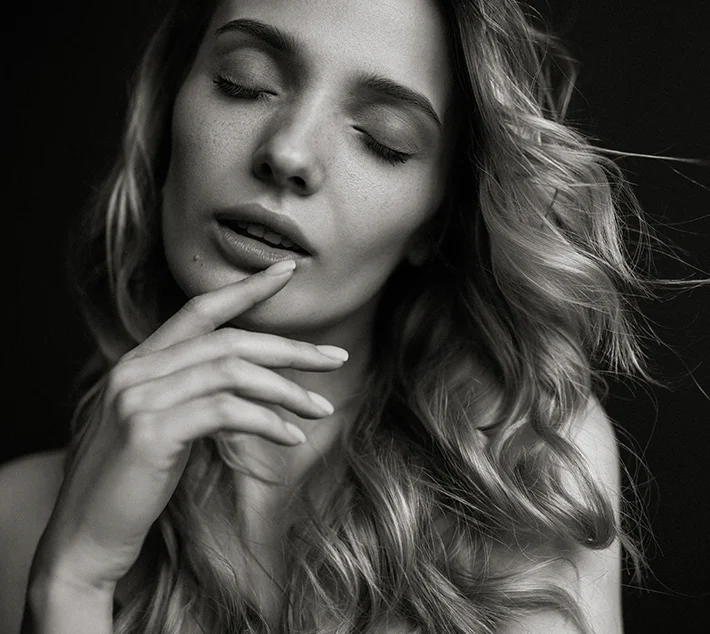A close-up of a woman with long, wavy hair, eyes closed, gently touching her lips, set against a dark background. - Youth Lift in West Village, New York, NY