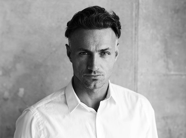 Black-and-white portrait of a man with short, styled hair, wearing a white shirt, against a textured gray background. - Men's Facelift in West Village, New York, NY