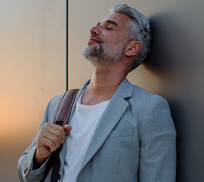 A relaxed man with gray hair and a beard leans against a wall, eyes closed, holding a bag, exuding calmness at sunset. - Men's Facelift in West Village, New York, NY