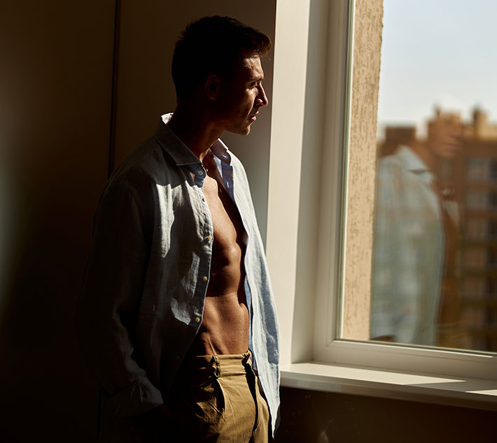 A shirtless man stands by a window, gazing thoughtfully outside, with sunlight casting warm tones on his figure. - Men's High Definition Liposutcion in West Village, New York, NY