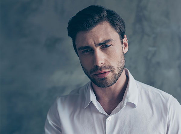 A thoughtful young man with short, styled hair and a beard, wearing a white shirt against a muted gray background. - Men's Jawline Contouring in West Village, New York, NY