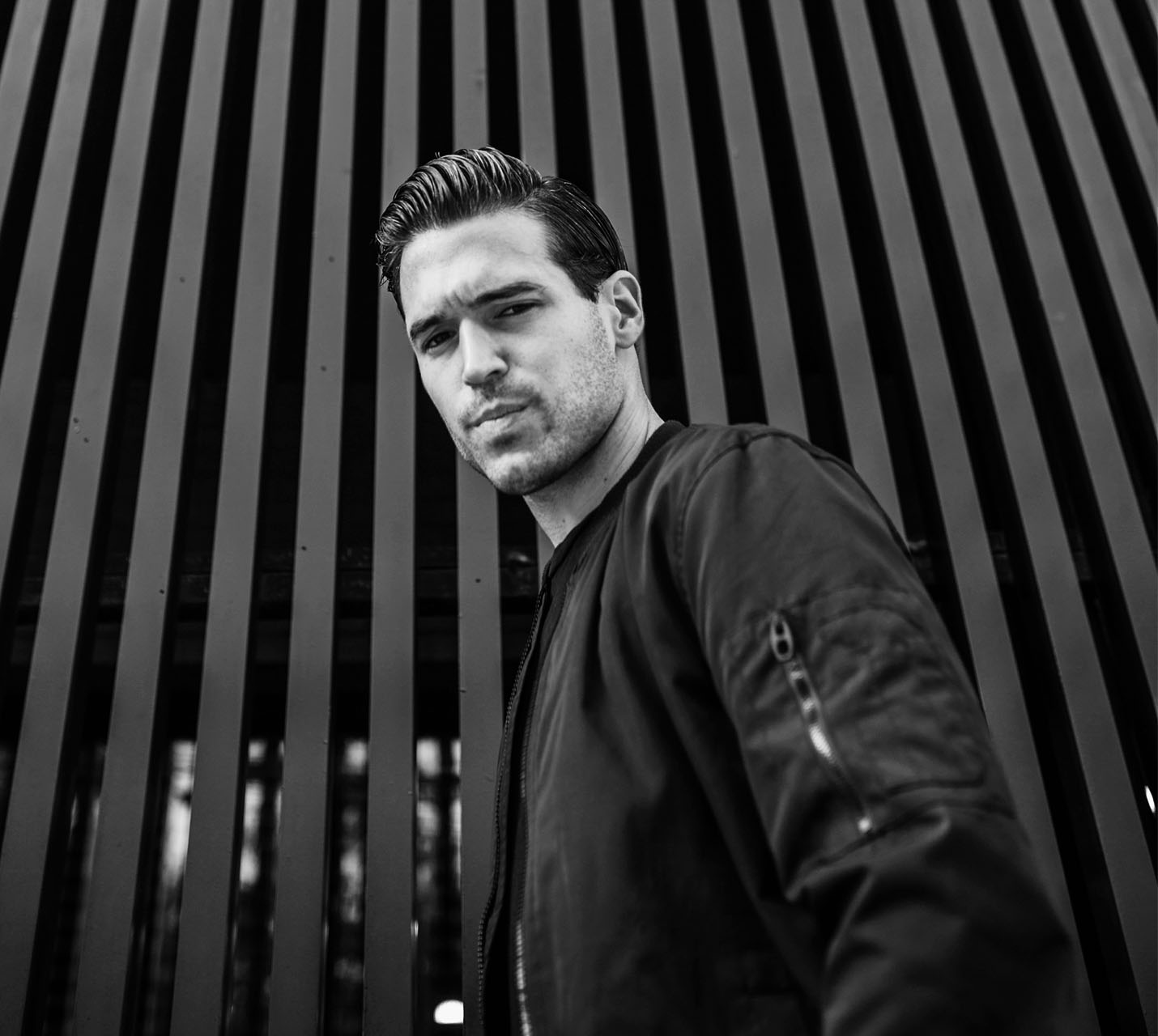A man poses confidently against a backdrop of vertical black slats, wearing a dark bomber jacket and showcasing slicked-back hair. - Men's Jawline Contouring in West Village, New York, NY