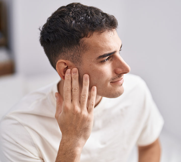 A young man with short dark hair is gently pressing his hand against his ear, listening attentively. He wears a light-colored shirt. - Men's Otoplasty in West Village, New York, NY