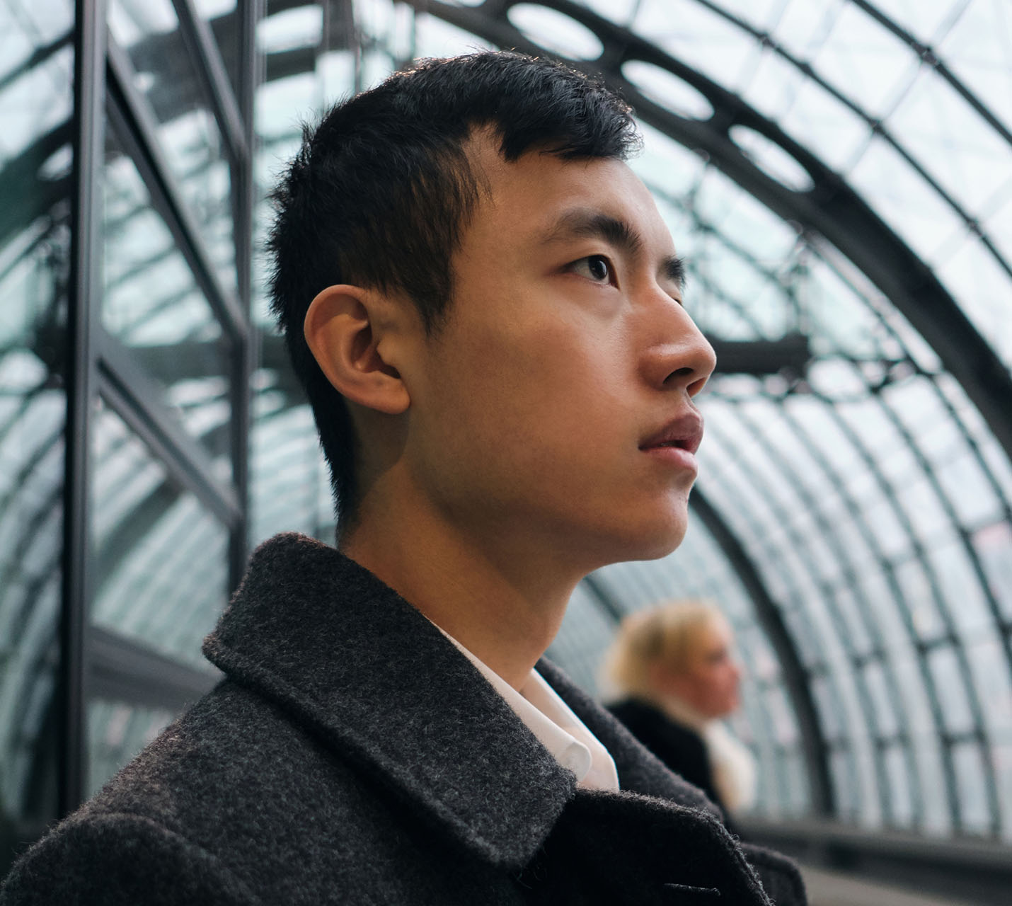 A young man in a gray coat looks thoughtfully to the side, with a modern glass arch in the background. A woman is blurred behind him. - Men's Otoplasty in West Village, New York, NY