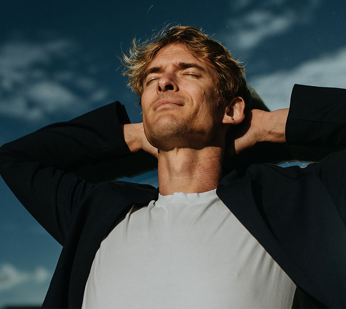 A young man with tousled blonde hair relaxes outdoors, arms behind his head, eyes closed, enjoying the sunny sky. - Men's Necklift in West Village, New York, NY