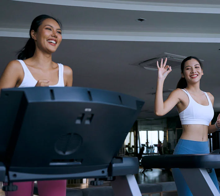 Two smiling women jog on treadmills in a bright gym, exuding energy and enjoyment in their workout. - Breast Reduction in West Village, New York, NY