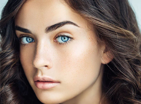 Close-up of a young woman with long, wavy hair, striking blue eyes, and a natural complexion featuring light freckles. - Eyelid Surgery in West Village, New York, NY