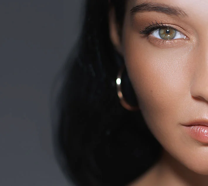Close-up of a young woman with striking green eyes, smooth skin, and dark hair, wearing a hoop earring against a dark background. - Fat Grafting in West Village, New York, NY