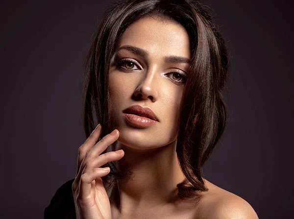 A close-up of a young woman with long, dark hair, soft features, and a natural makeup look, gently touching her face. - Rhinoplasty in West Village, New York, NY