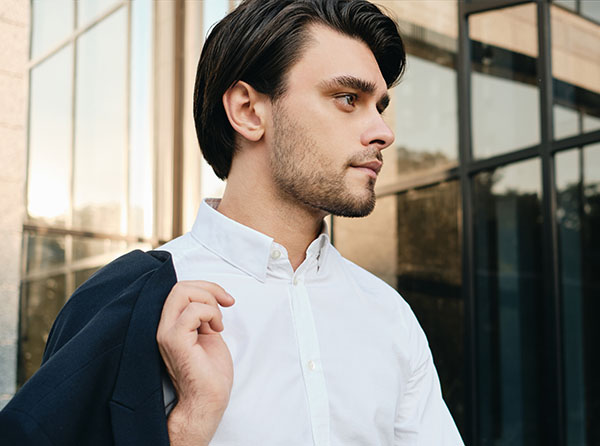 A young man in a white shirt holds a dark blazer over his shoulder, gazing thoughtfully in an urban setting. - Men's Rhinoplasty in West Village, New York, NY