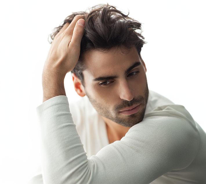 A young man with tousled dark hair gazes thoughtfully to the side, resting his hand on his head, wearing a light sweater. - Men's Rhinoplasty in West Village, New York, NY