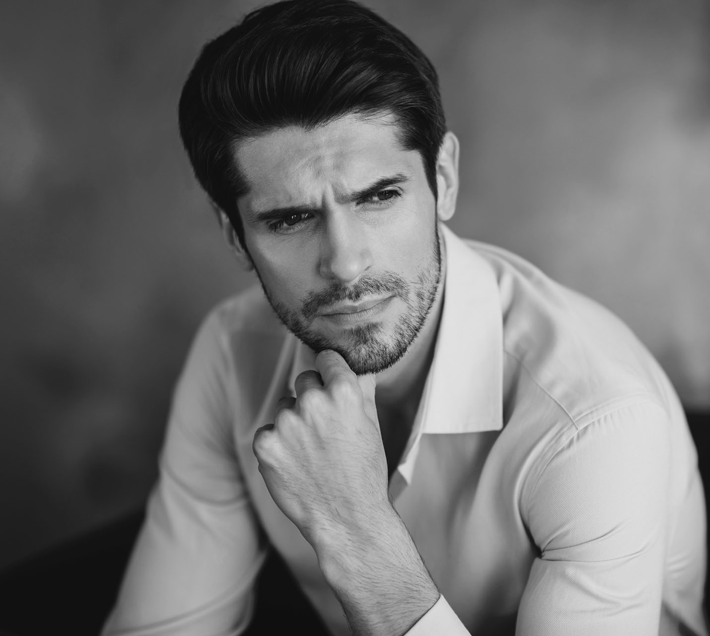 A man with a thoughtful expression rests his chin on his hand, wearing a light shirt against a muted background. - The Difference with Male Facial Surgery in West Village, New York, NY