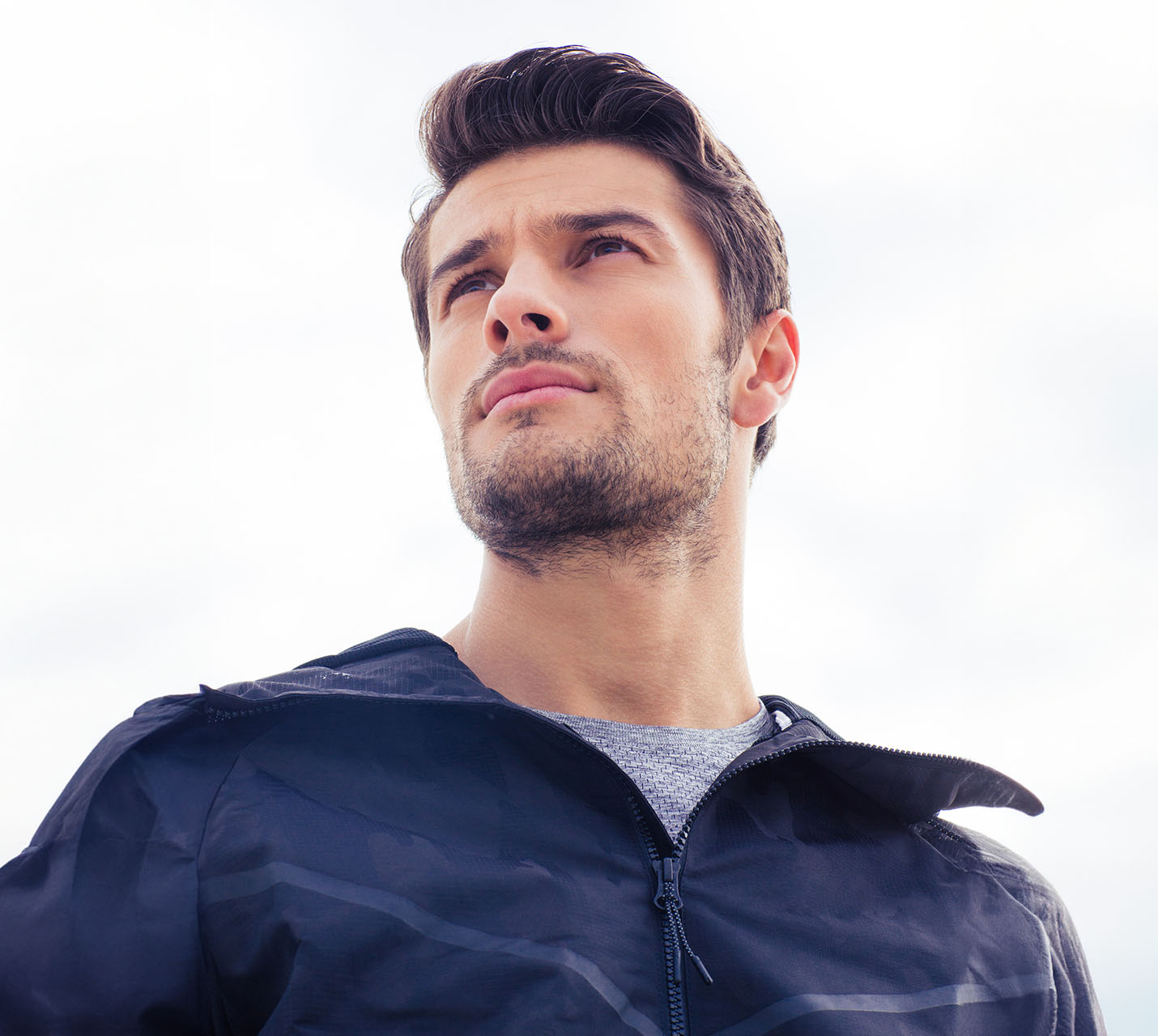 A young man in a black jacket gazes upward, with a confident expression against a cloudy sky background. - The Difference with Male Facial Surgery in West Village, New York, NY