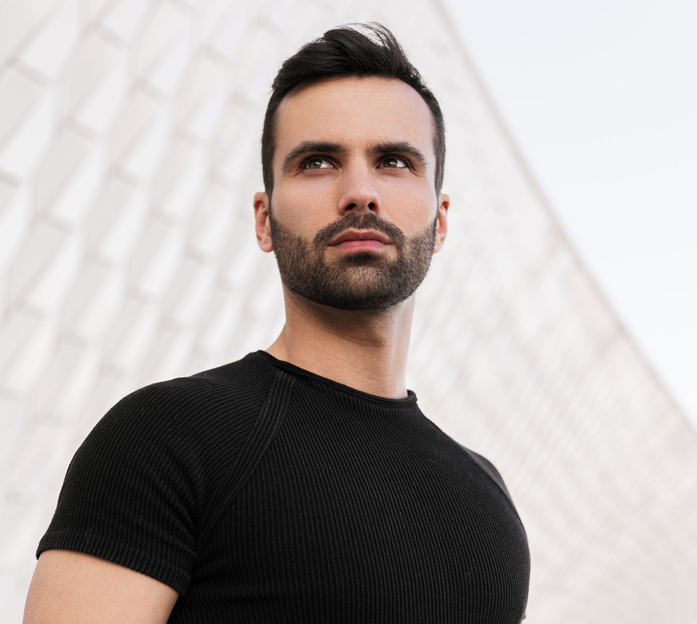 A man with a beard and short black hair looks thoughtfully into the distance, wearing a black ribbed t-shirt against a light background. - The Male Aesthetic in West Village, New York, NY
