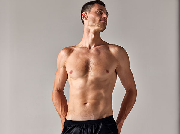 A muscular man stands shirtless against a light gray background, looking slightly upwards with a confident posture. - Mens Tummy Tuck in West Village, New York, NY